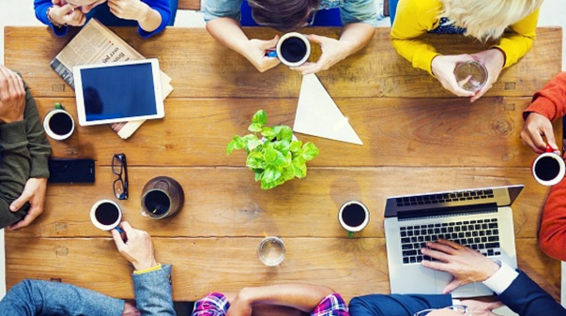 Eine Gruppe Menschen sitzt mit Laptops und Kaffee an einem Tisch und hat ein Meeting.