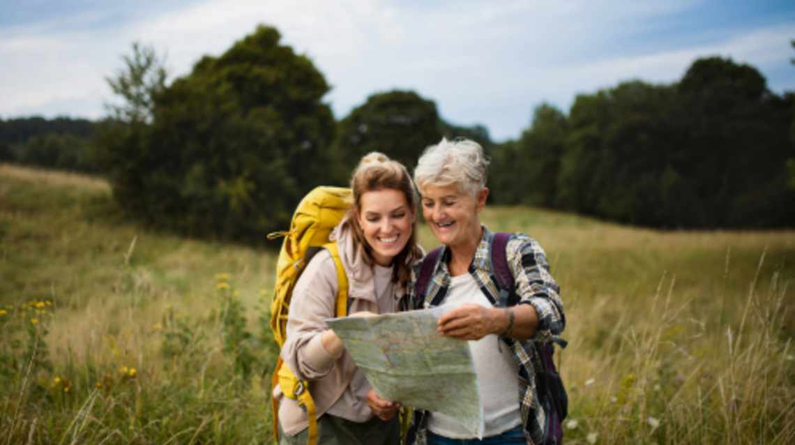 Eine jüngere und eine ältere Frau schauen beim Wandern in schöner Landschaft auf eine Karte.